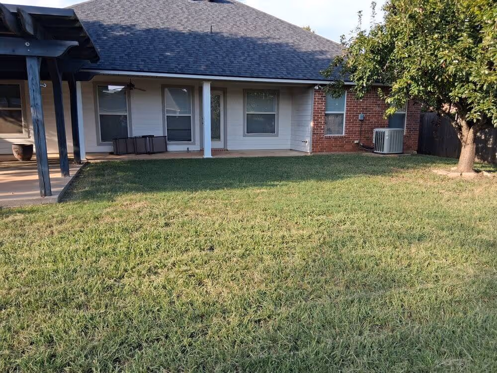 Backyard with a neatly mowed lawn, a tree on the right, and a house with a covered patio and air conditioning unit.