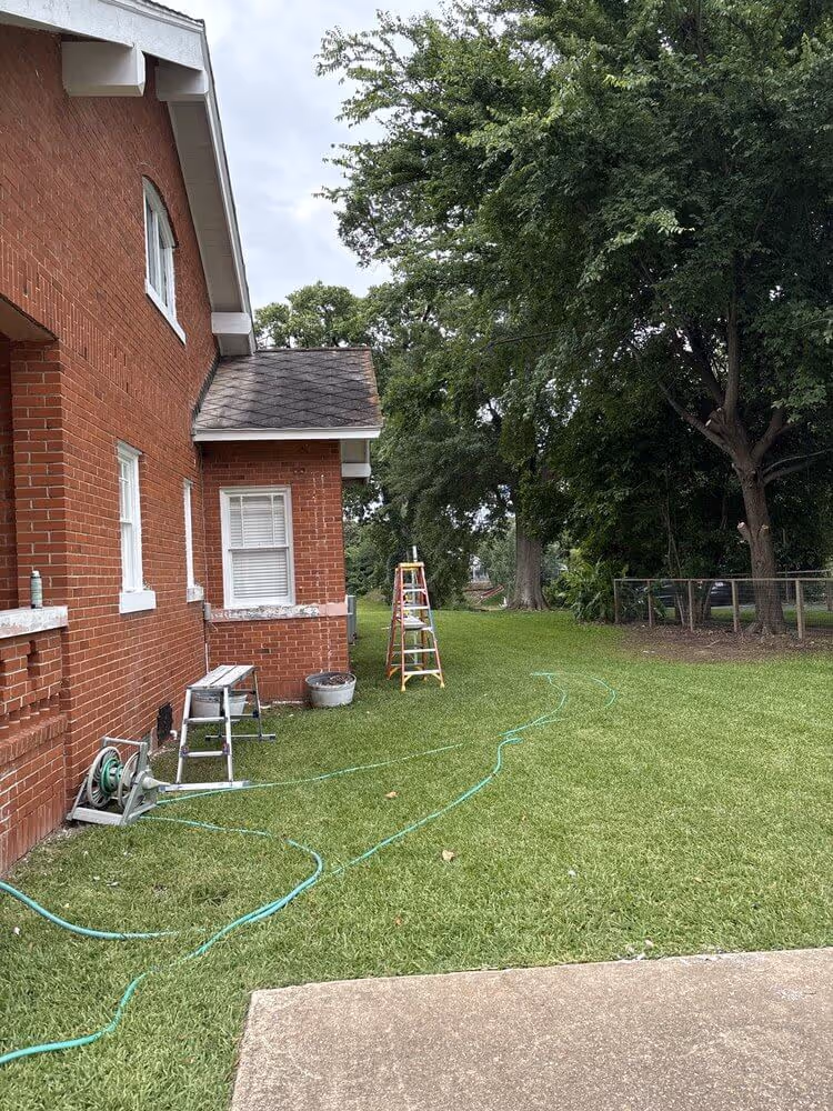 Side yard of a red brick house with green grass, a garden hose, a stepladder, and large trees in the background.